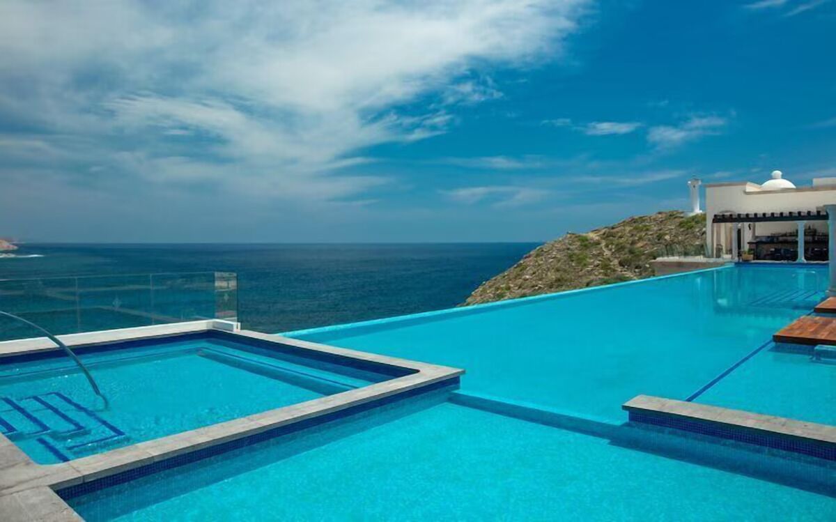 A clear blue infinity pool overlooking the ocean, with a white building and a small hill in the background.
