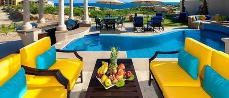 A poolside area with yellow and blue outdoor furniture, a table with fruit, and a view of the ocean.
