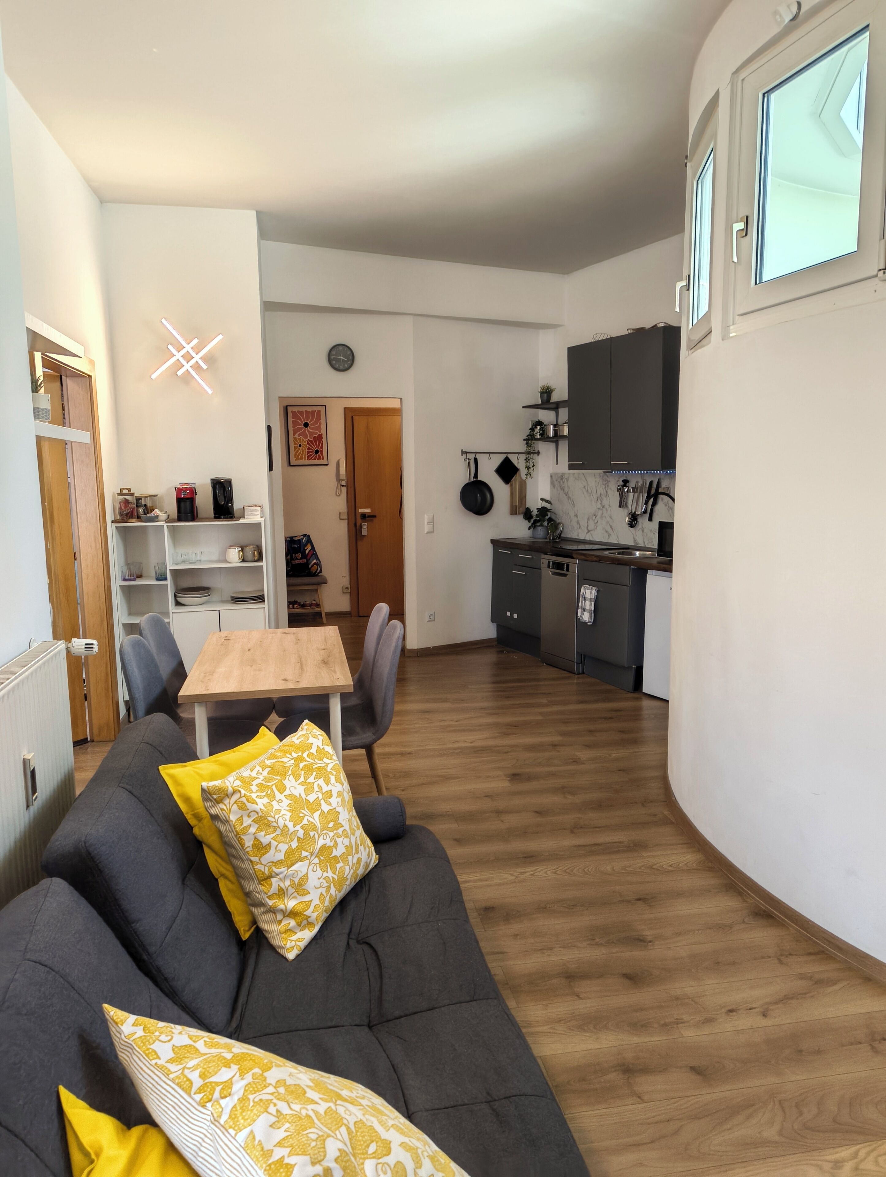 A modern living room with a sofa, a dining table, and chairs. There is a kitchen area with cabinets and a countertop.