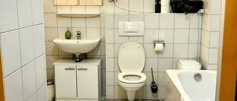 Un baño con lavamanos, inodoro y bañera blancos. Hay un espejo sobre el lavamanos y una repisa con plantas. El suelo tiene un diseño a cuadros.