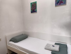 A single-bed room with a white mattress, a green pillow, and a folded towel. There is a small painting on the wall above the bed.