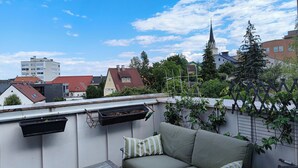 Una terraza en la azotea con un sofá y una mesa, con vistas a un paisaje urbano con edificios y una aguja de iglesia.