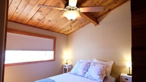 Un dormitorio con techo de madera, cama con ropa de cama floral, dos mesitas de noche con lámparas y una ventana con persianas.