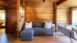 Interior acogedor de una cabaña de troncos con un sofá seccional gris, vigas de madera y una mesa de comedor de madera.