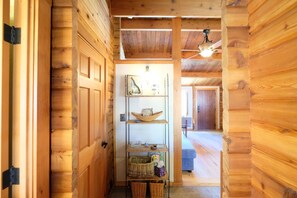 Un couloir intérieur aux murs en bois, avec une étagère en métal où sont exposés divers objets, un ventilateur de plafond et une porte donnant sur une autre pièce.