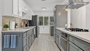 A modern kitchen with grey cabinets, a stainless steel range hood, and a window with blinds. - Traveler's Deam Home - SDSU - Hot Tub - Pool Table (San Diego)
