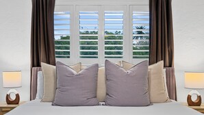 A bedroom with a large bed, two bedside tables with lamps, and a view of greenery through the window. - Traveler's Deam Home - SDSU - Hot Tub - Pool Table (San Diego)