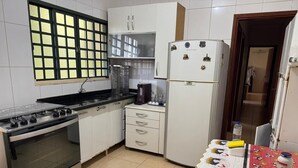 A compact kitchen with a gas stove, oven, white cabinets, a refrigerator, and a table with a patterned tablecloth. - ACCOMMODATION BARRETOS PEÃO FESTIVAL - 2025 (América Barretos)