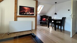Un salon moderne avec un plancher en bois, un radiateur blanc, un téléviseur fixé sur une poutre en bois, une table à manger avec des chaises et une lucarne.