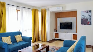 Ein Wohnzimmer mit zwei blauen Sofas, einem Couchtisch, einem Fernseher auf einer Holzkommode und gelben Vorhängen.
