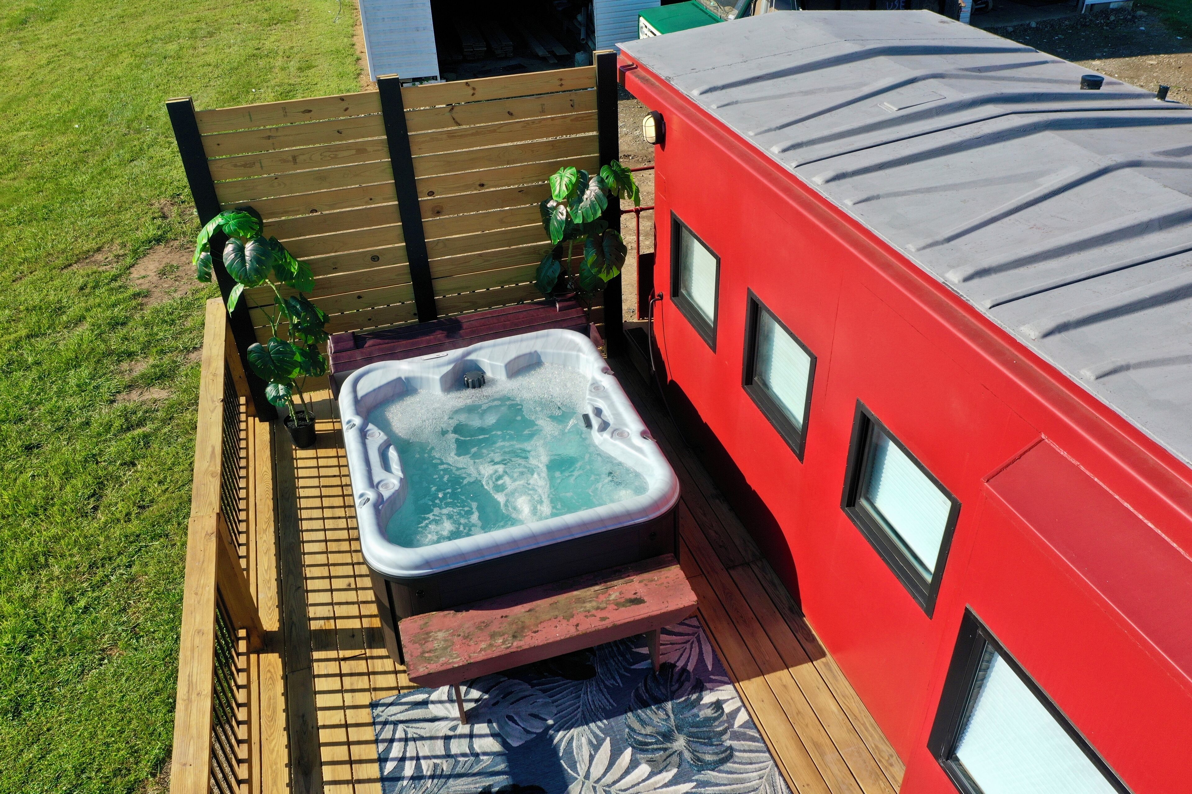 A hot tub adjacent to a red building with windows, surrounded by a wooden deck and a fence.