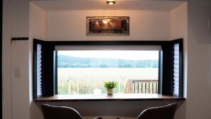 Quarto com uma janela grande, duas cadeiras, um vaso de planta e um quadro na parede.