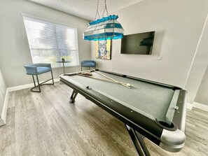 Una mesa de billar con tacos, una silla azul y un cuadro en la pared.