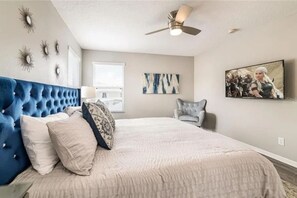 A bedroom with a tufted headboard, a bed with a textured cover, a ceiling fan, a window with blinds, a gray armchair, and a painting on the wall. - The Minions Mansion 6br Resort Pool Home By Disney (Kissimmee)