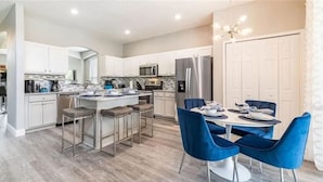A modern kitchen with a central island, bar stools, and a dining area with blue chairs. - The Minions Mansion 6br Resort Pool Home By Disney (Kissimmee)