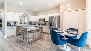 A modern kitchen with a central island, bar stools, and a dining area with blue chairs. - The Minions Mansion 6br Resort Pool Home By Disney (Kissimmee)