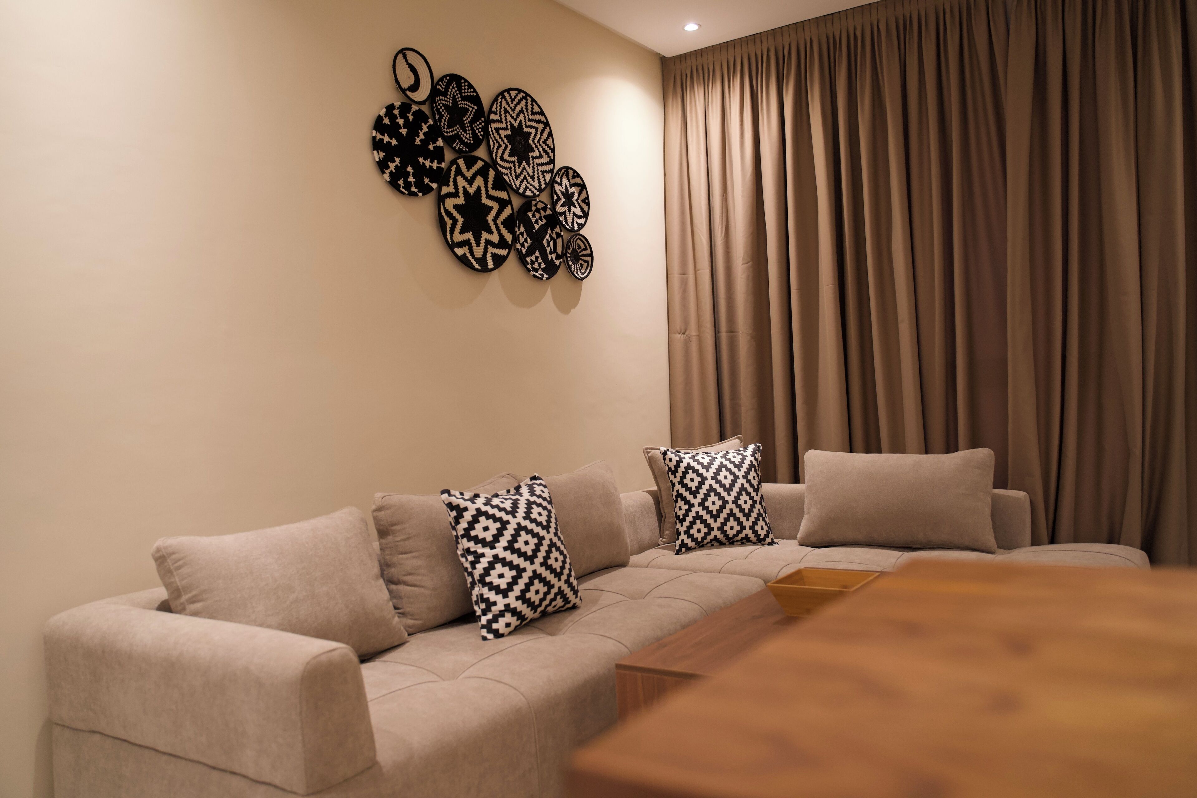 Uma sala de estar moderna com um sofá bege, almofadas estampadas, uma mesa de centro de madeira e um objeto decorativo fixado na parede.