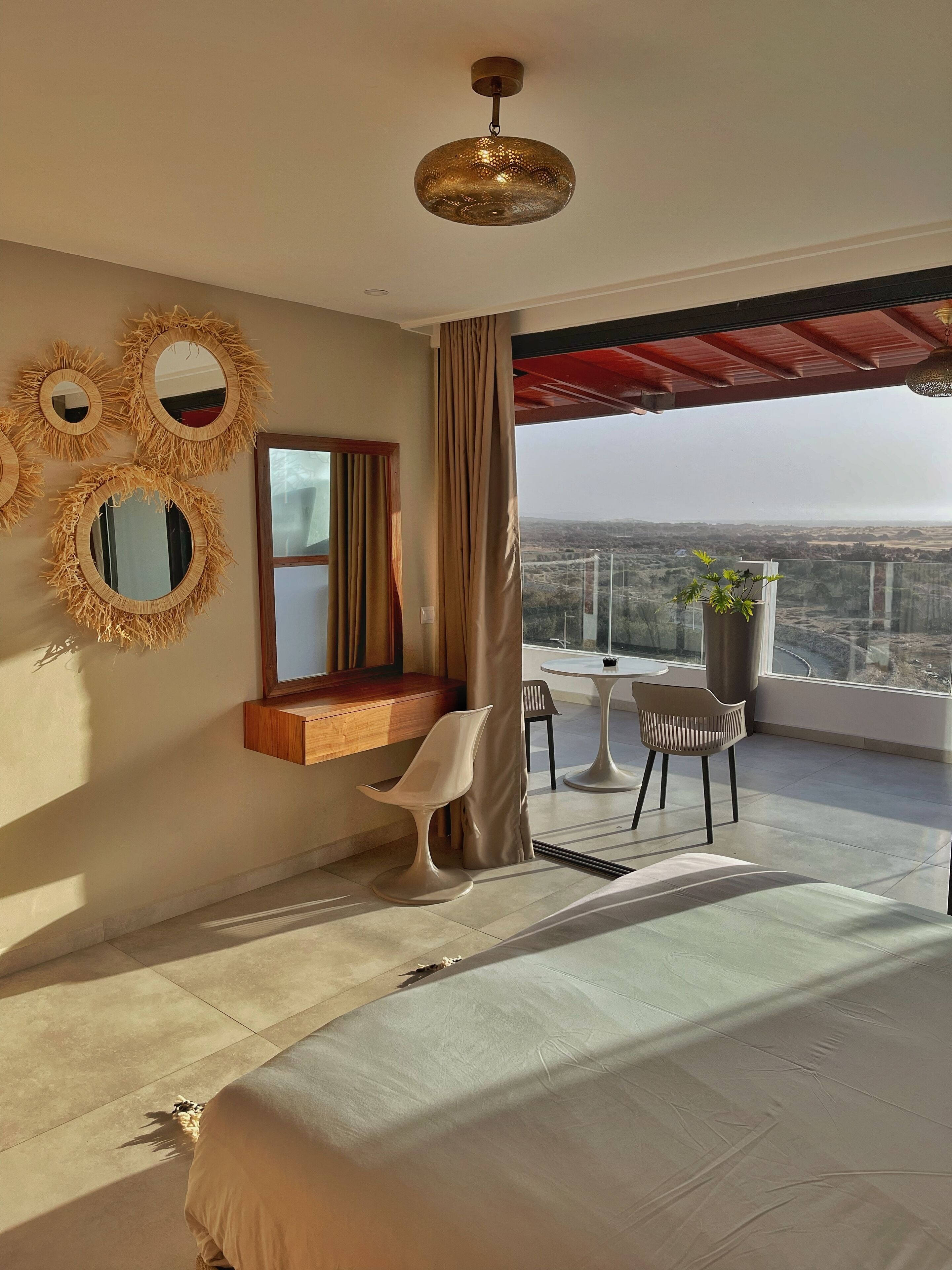 A modern bedroom with a large bed, a vanity with mirrors, a small table with chairs, and a balcony with a view.
