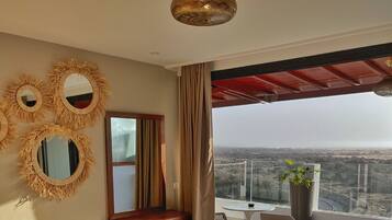 A modern bedroom with a large bed, a vanity with mirrors, a small table with chairs, and a balcony with a view.