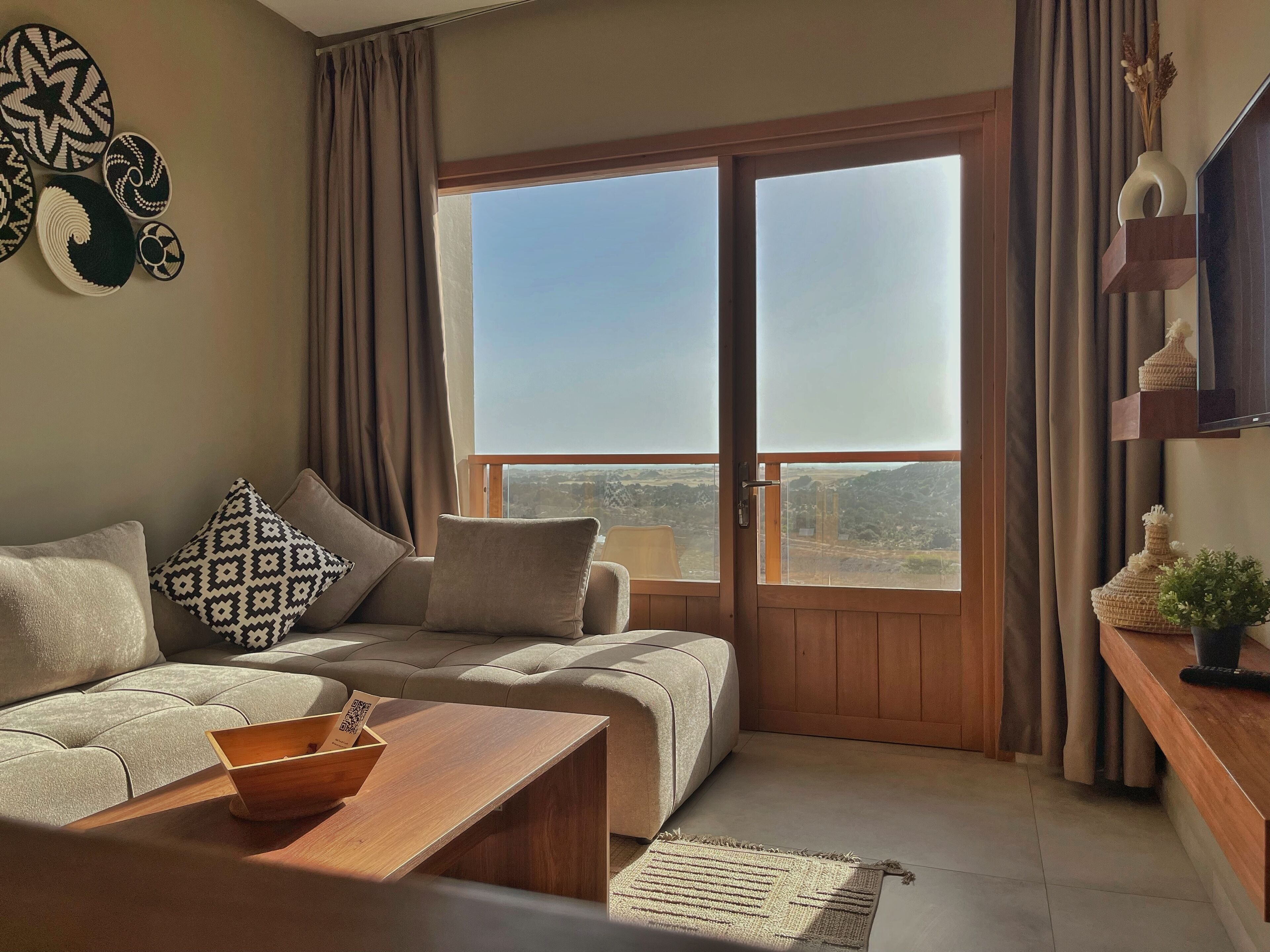 A modern living room with a sofa, a wooden coffee table, and a view of a mountainous landscape through a large sliding door.