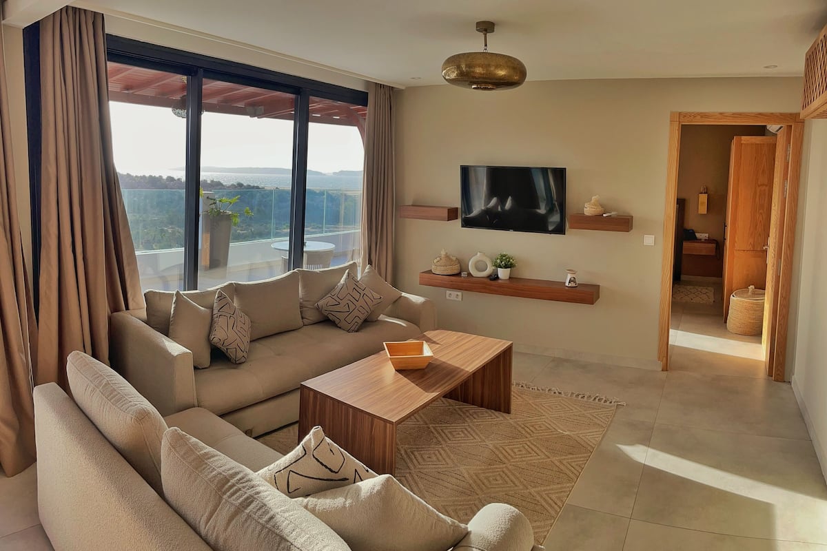 Een moderne woonkamer met een grote bank, een salontafel en een tv aan de muur bevestigd. Er is een balkon met uitzicht op de stad.
