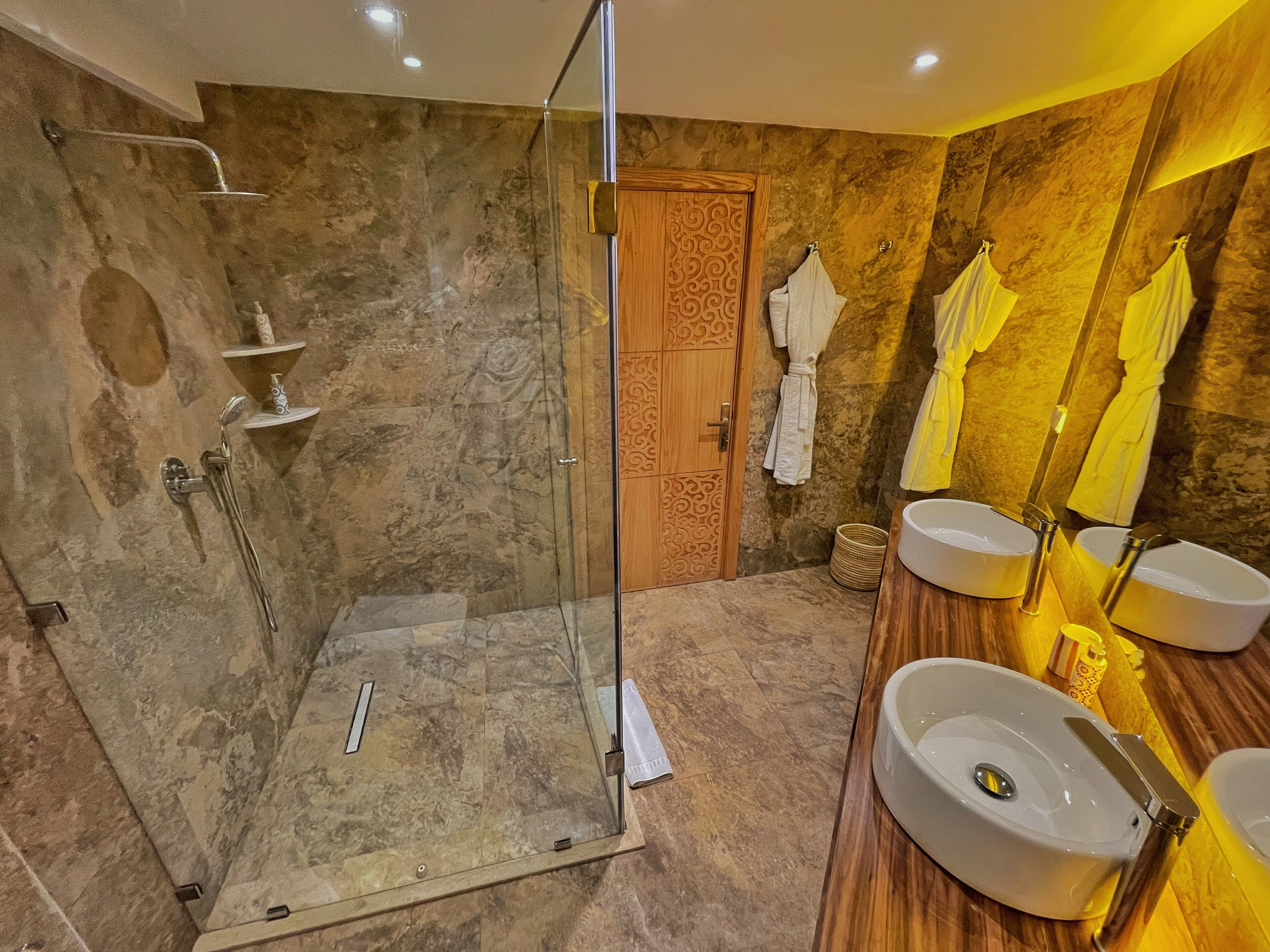 Un baño moderno con ducha de vidrio, paredes de mármol, puerta de madera y dos lavamanos con toallas amarillas.