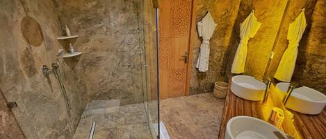 Un baño moderno con ducha de vidrio, paredes de mármol, puerta de madera y dos lavamanos con toallas amarillas.