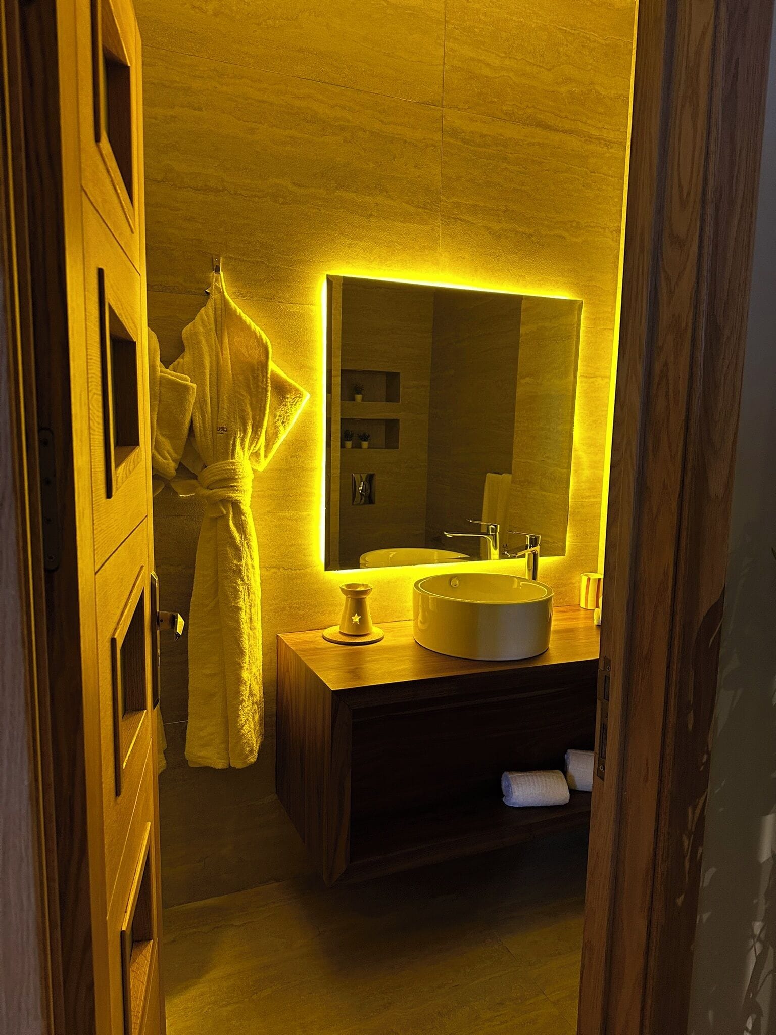 A bathroom with a wooden vanity, a large mirror, and a towel hanging on the door.