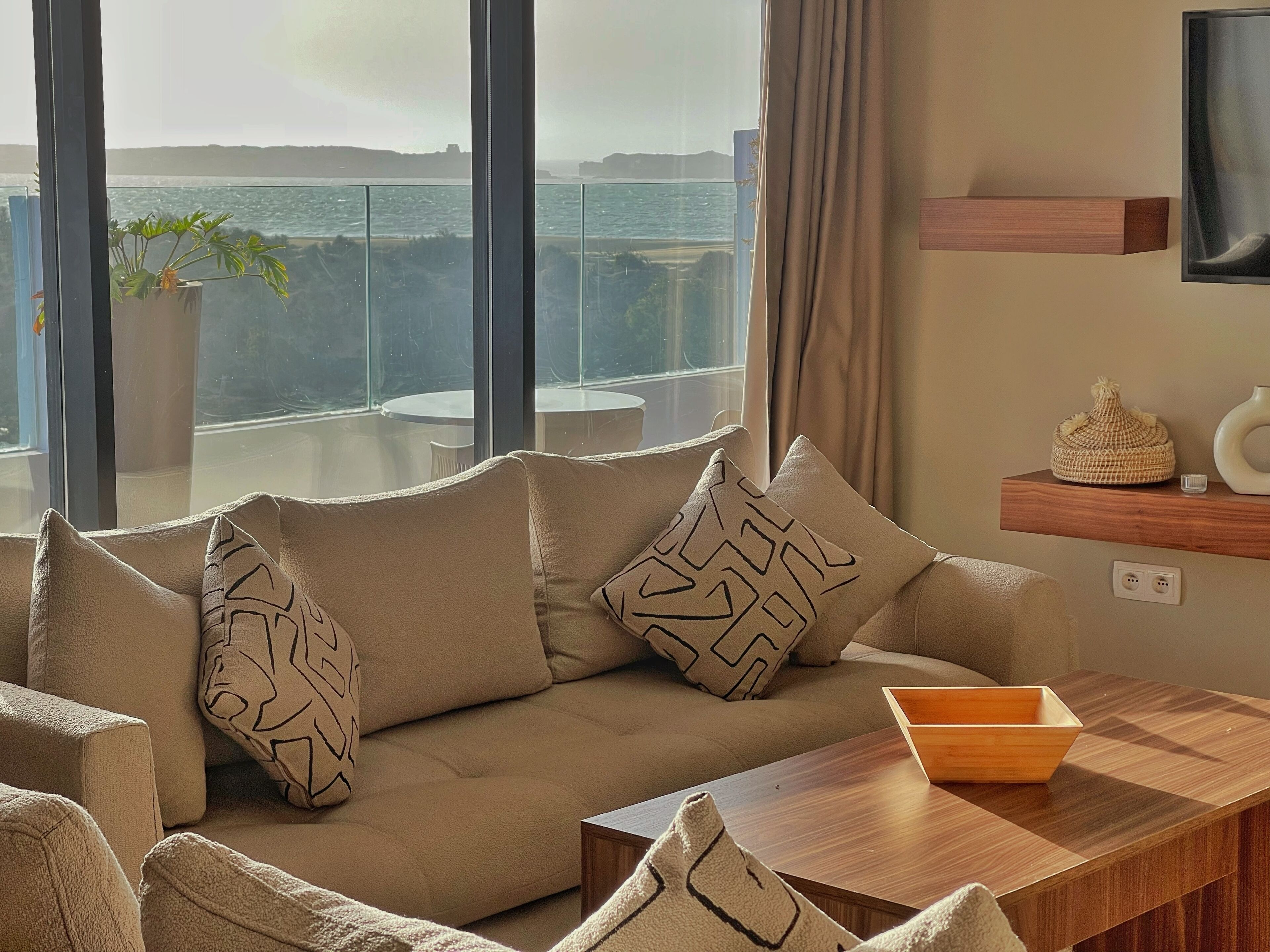 A modern living room with a beige sofa, patterned cushions, a wooden coffee table, and a view of the sea through large windows.