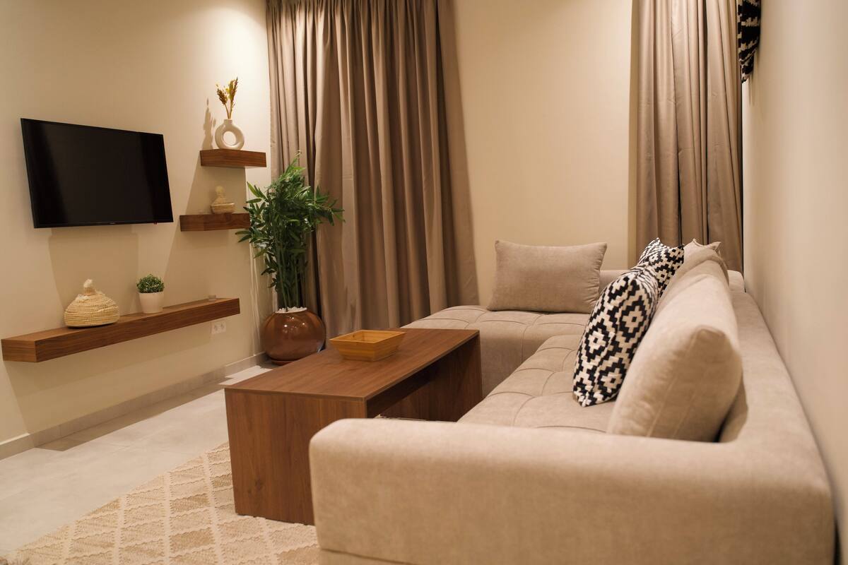 Een moderne woonkamer met een beige bank, een houten salontafel, een flatscreen televisie aan de muur en een kamerplant.