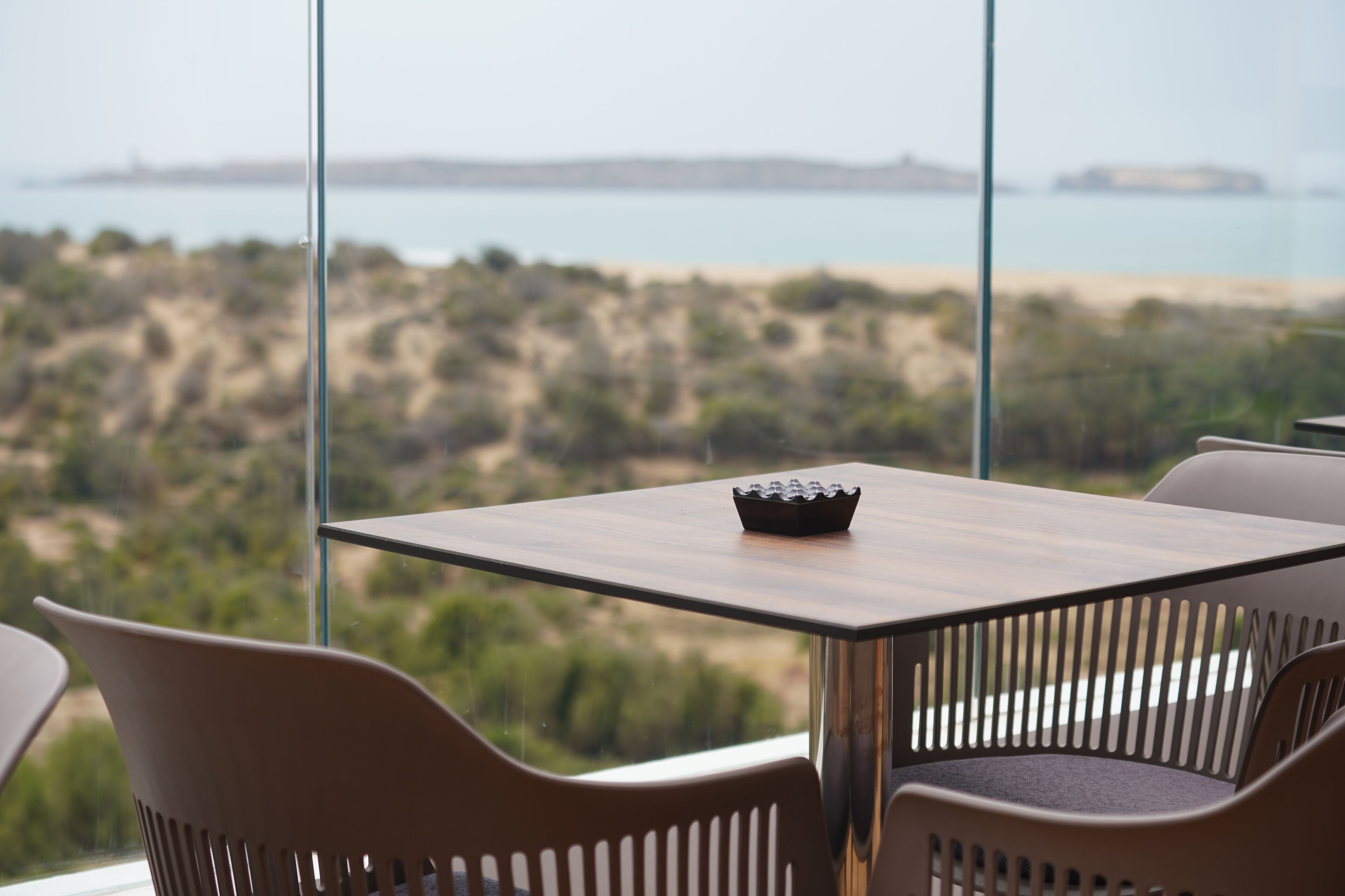 Una mesa con un bol, rodeada de sillas, y una vista a una playa y tierra firme distante a través de amplios ventanales.