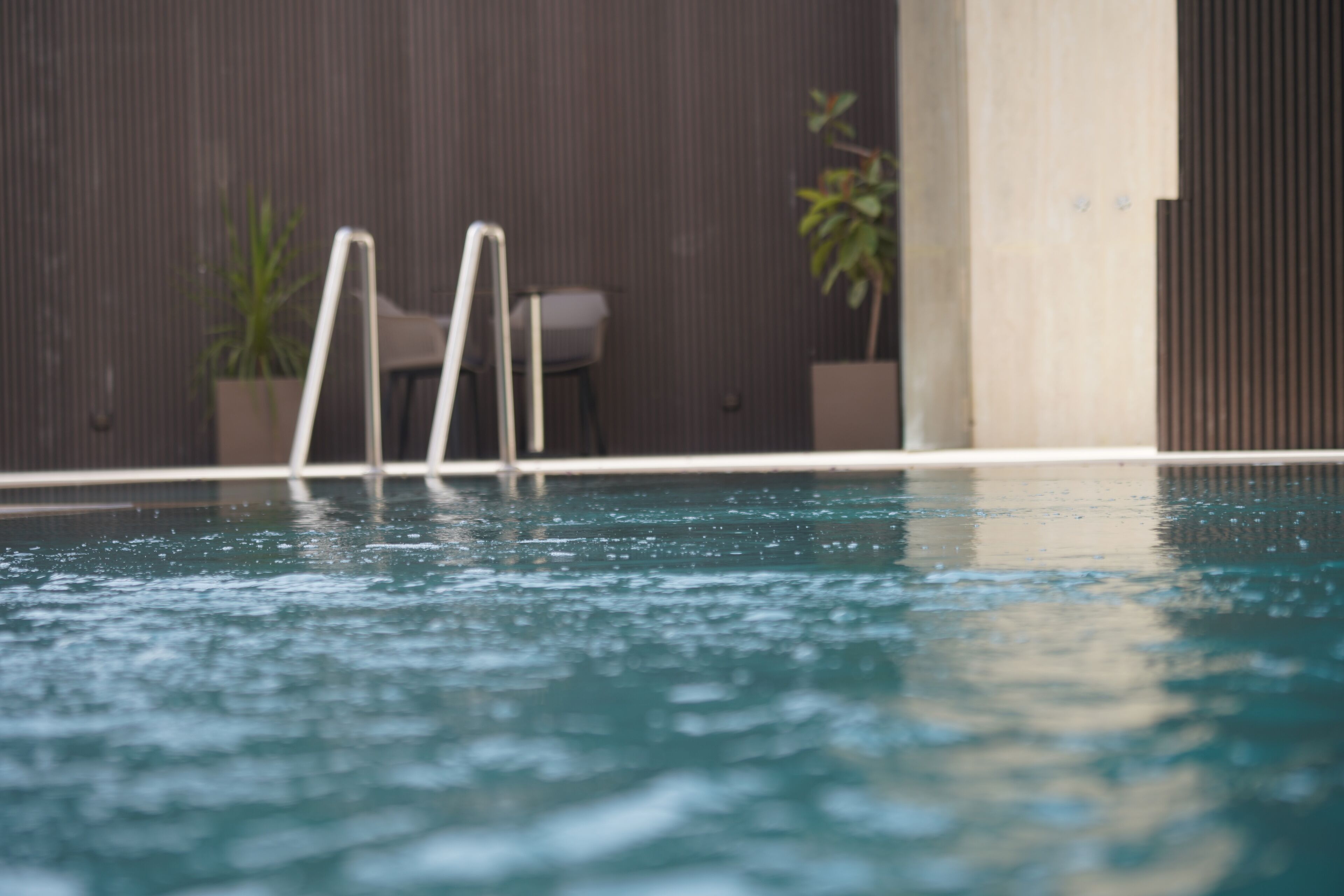 A swimming pool with a metal ladder, a chair, and potted plants against a textured wall.