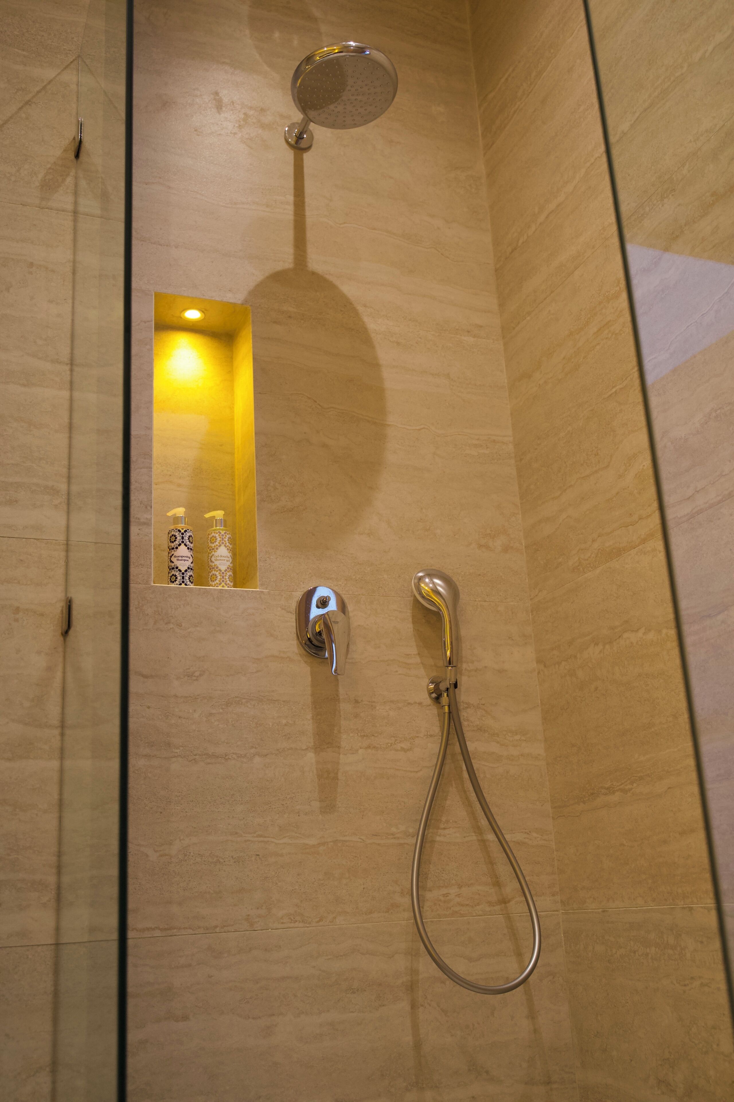 A shower with a handheld showerhead and a wall-mounted showerhead, a small mirror with a light, and two bottles of shampoo and conditioner.