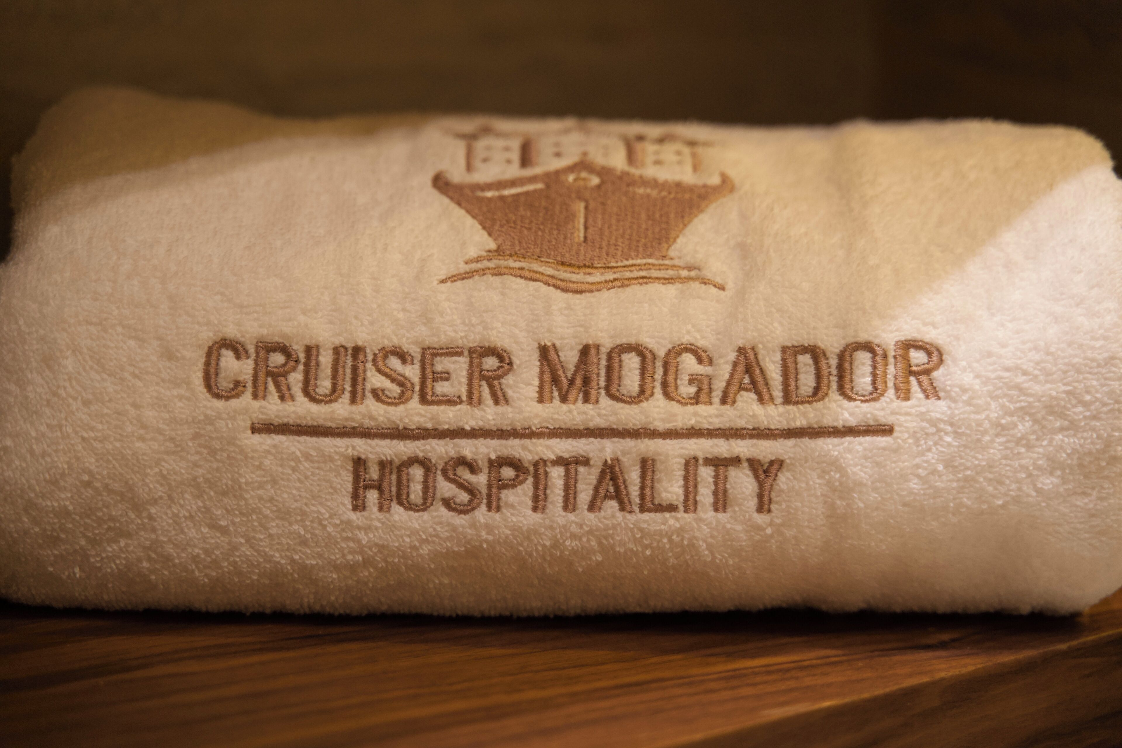 Una toalla blanca con bordado que dice 'CRUISER MOGADOR HOSPITALITY' y un emblema de corona.