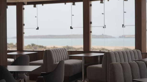 A modern interior with seating and tables arranged near large windows overlooking a beach. - Cruiser Mogador Hospitality (Essaouira)