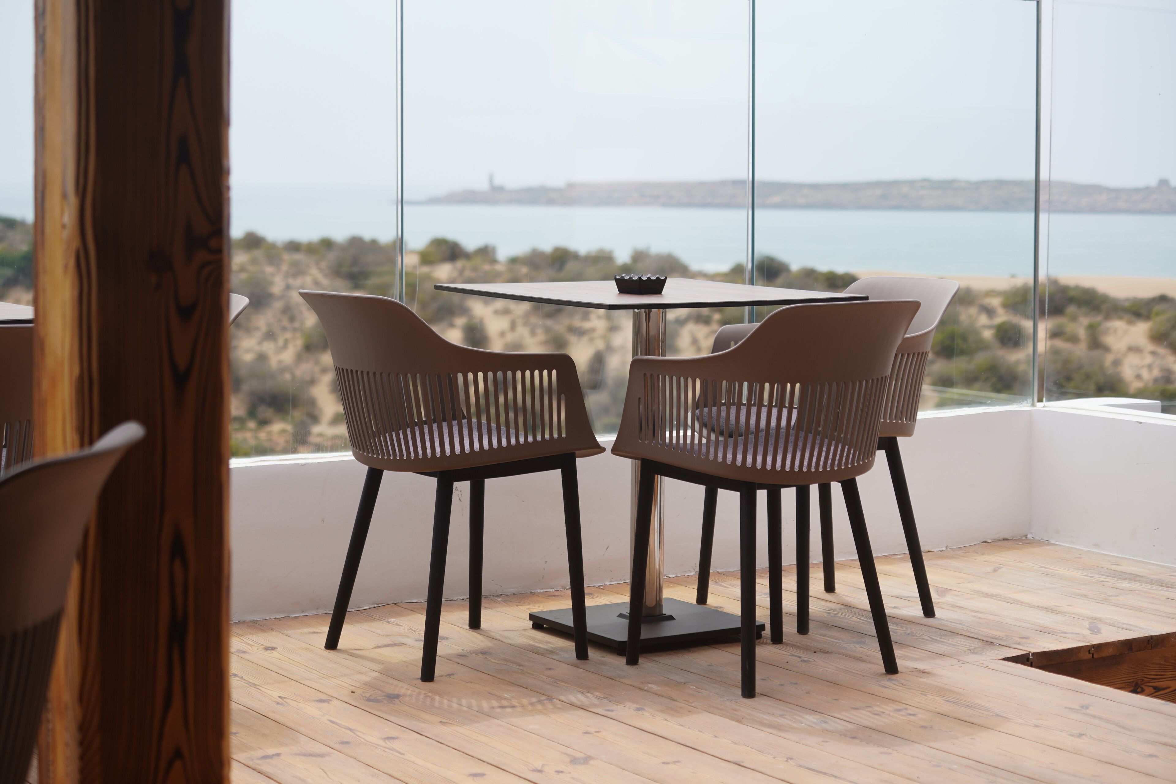 A modern dining area with a table and two chairs, large windows with a view of the sea and hills.