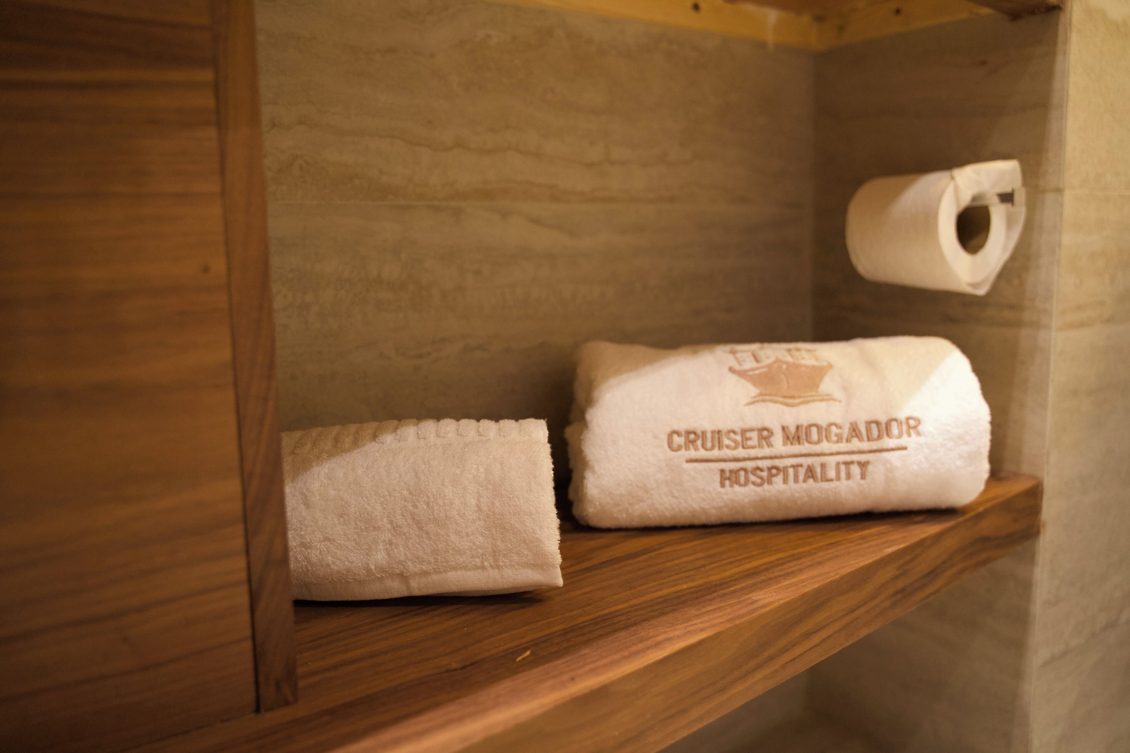 A wooden shelf with a white towel and a branded towel from 'Cruiser Mogador Hospitality'.