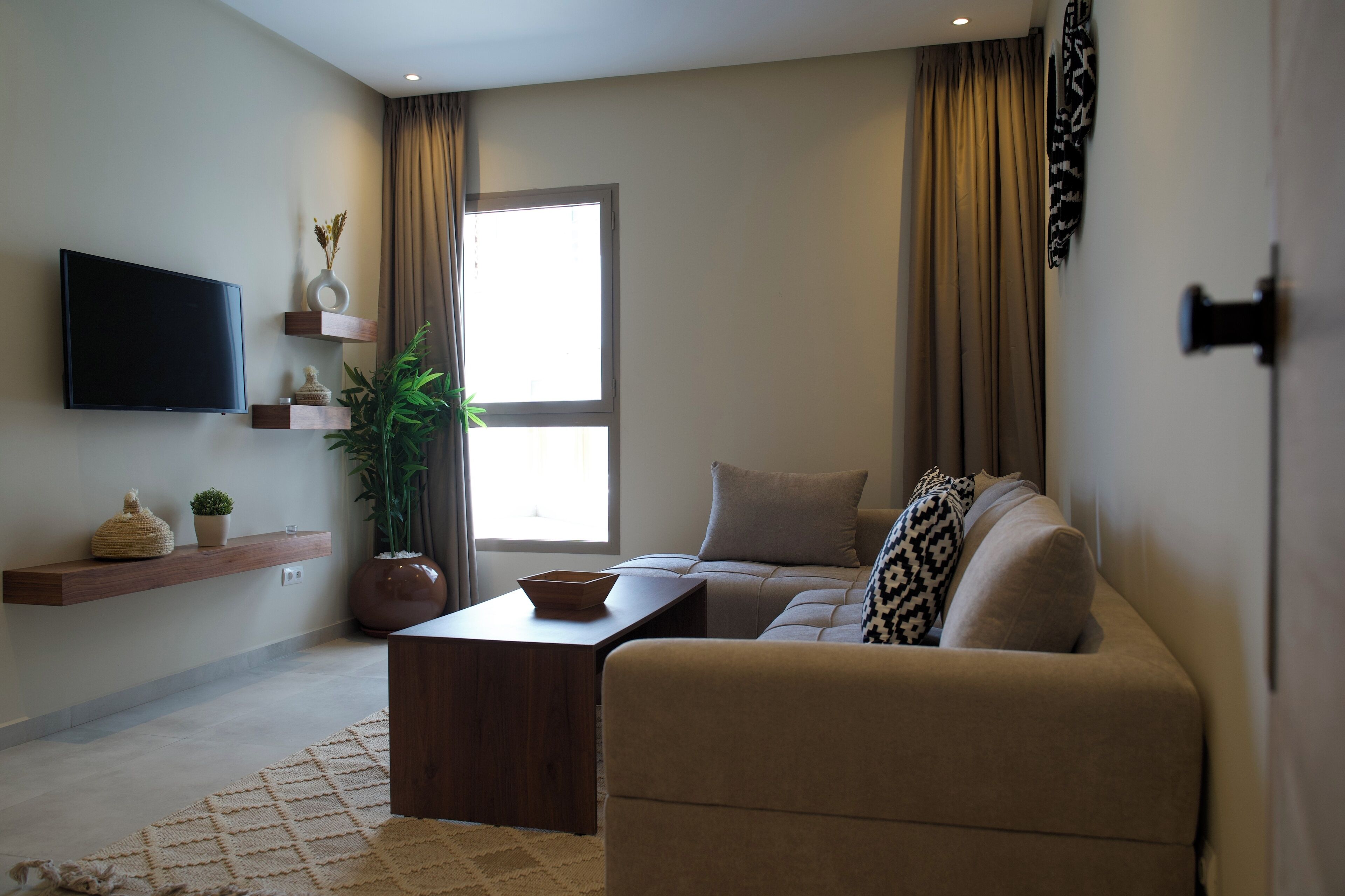 A modern living room with a flat-screen TV, wooden shelves, a sofa, a coffee table, and a large window with curtains.