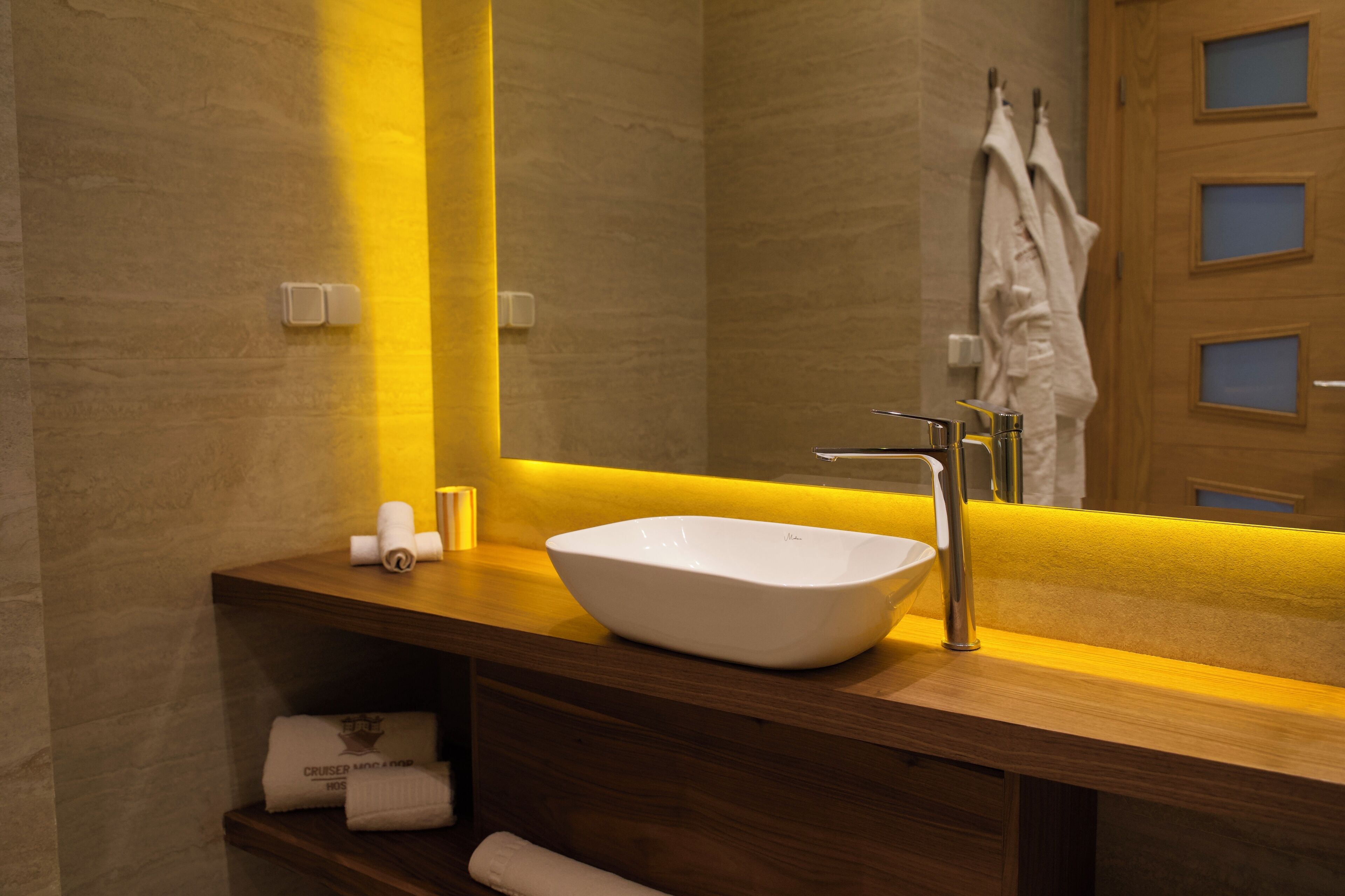 Un baño moderno con tocador de madera, lavamanos tipo cuenco blanco y un espejo que refleja un perchero y una puerta.