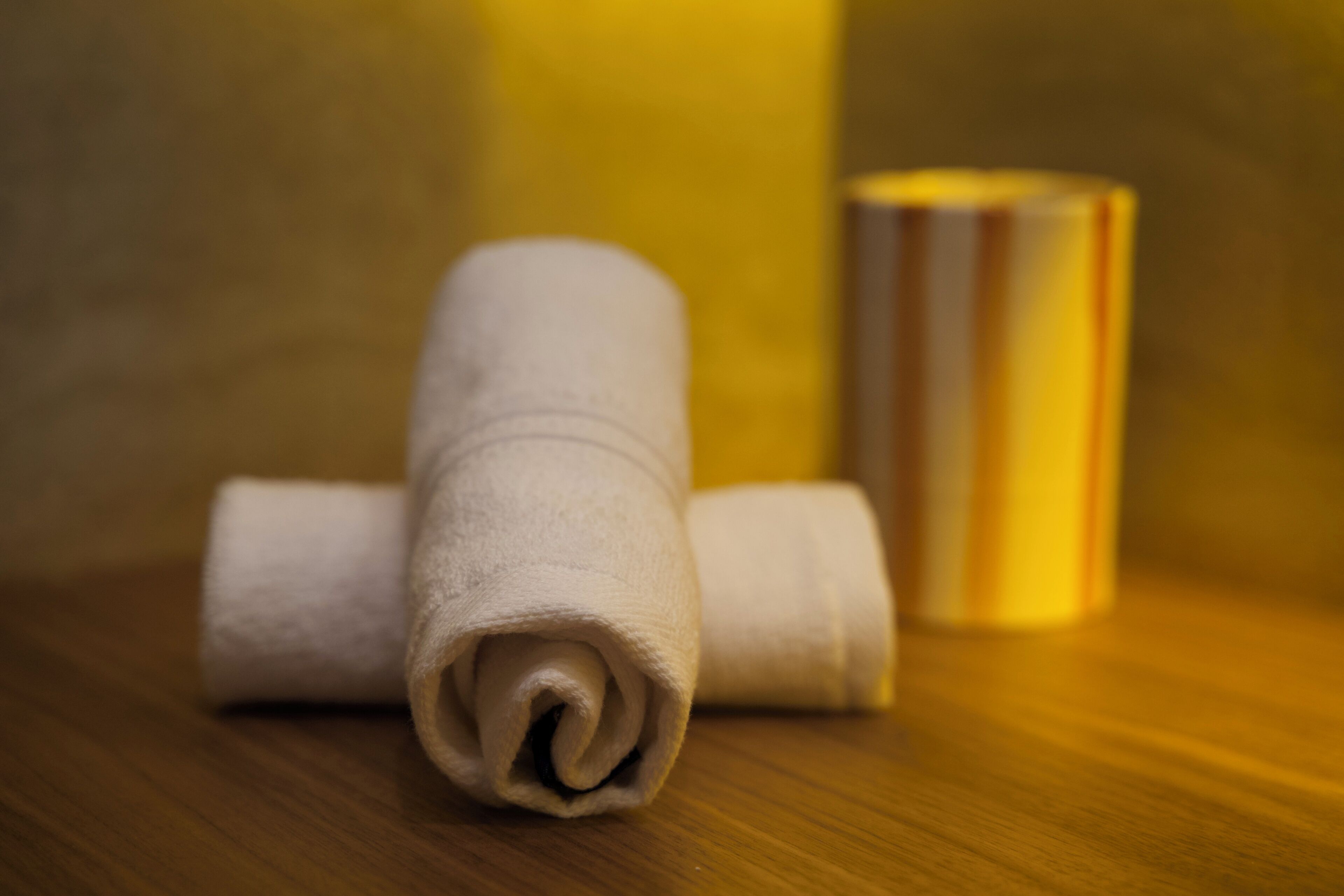 A rolled white towel on a wooden surface with a cylindrical object in the background.