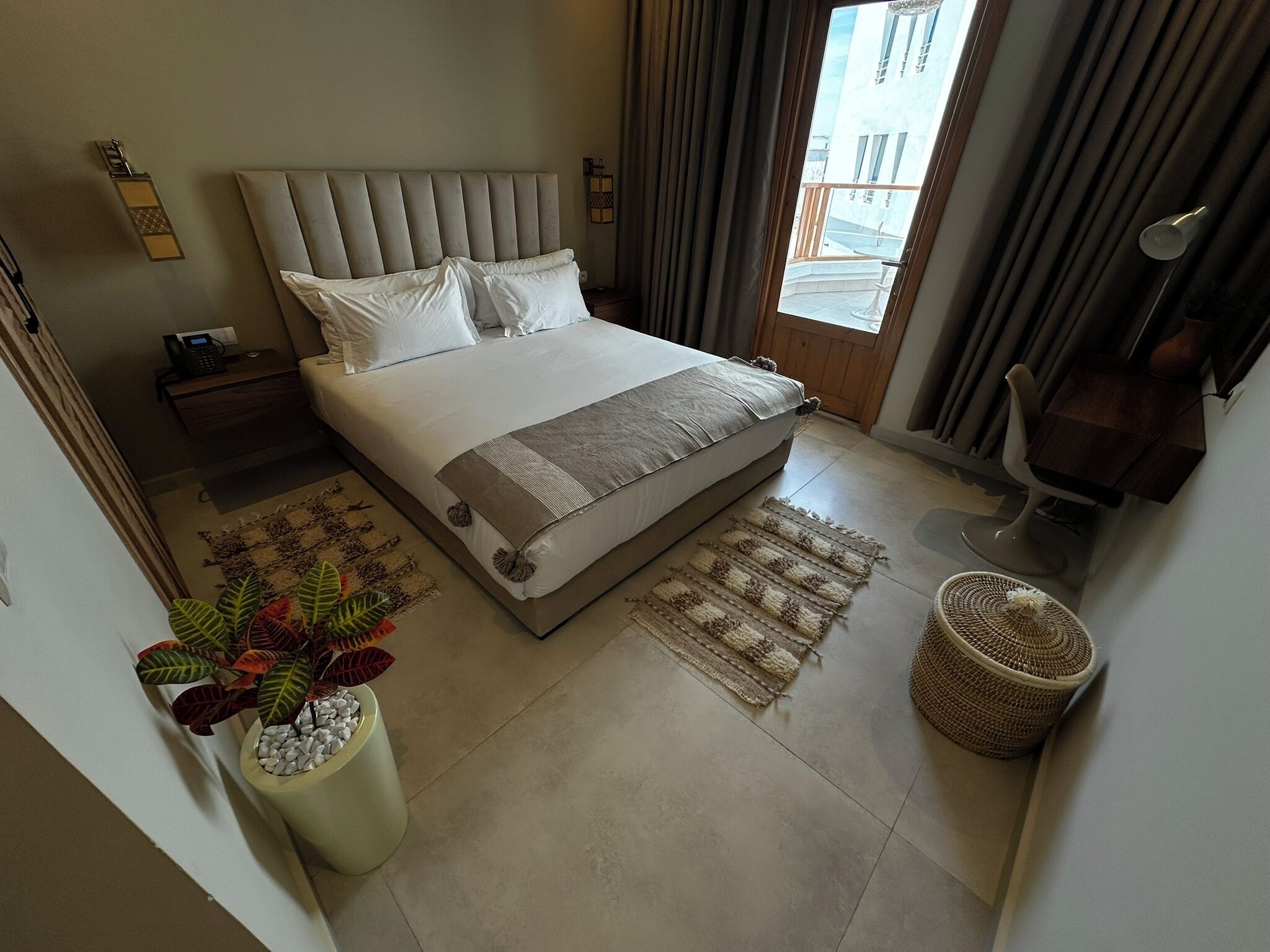 Quarto moderno com uma cama grande, mesa de cabeceira com telefone, planta em vaso, tapete tecido e vista para a varanda externa.