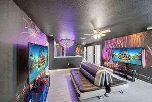 Un salon moderne comprenant un canapé, deux téléviseurs à écran plat, un ventilateur de plafond et un mur orné d’une fresque colorée.