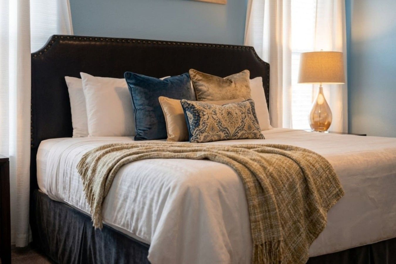 Un lit avec une tête de lit sombre, plusieurs oreillers, un plaid et une lampe de chevet.