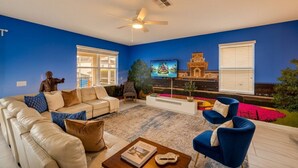 A living room with a beige sofa, blue armchairs, a coffee table, a TV, and a ceiling fan. - The Magic Kingdom 9BR Resort Pool Home Near Disney (Four Corners)