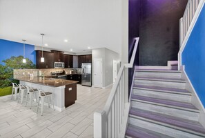 A modern kitchen with a central island, bar stools, and a staircase with a purple railing. - The Magic Kingdom 9BR Resort Pool Home Near Disney (Four Corners)