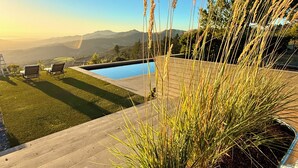 A pool with a view of mountains, a grassy area with two chairs, and tall grasses in the foreground.