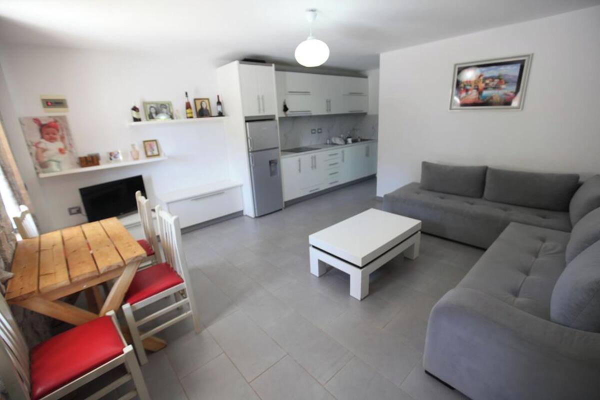 A living room with a grey sofa, a white coffee table, and a wooden dining table with red chairs. There is a kitchen area with white cabinets and a white ceiling light.