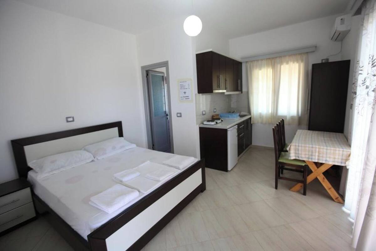 A bedroom with a bed, a small kitchenette, a dining table with chairs, and a window with curtains.