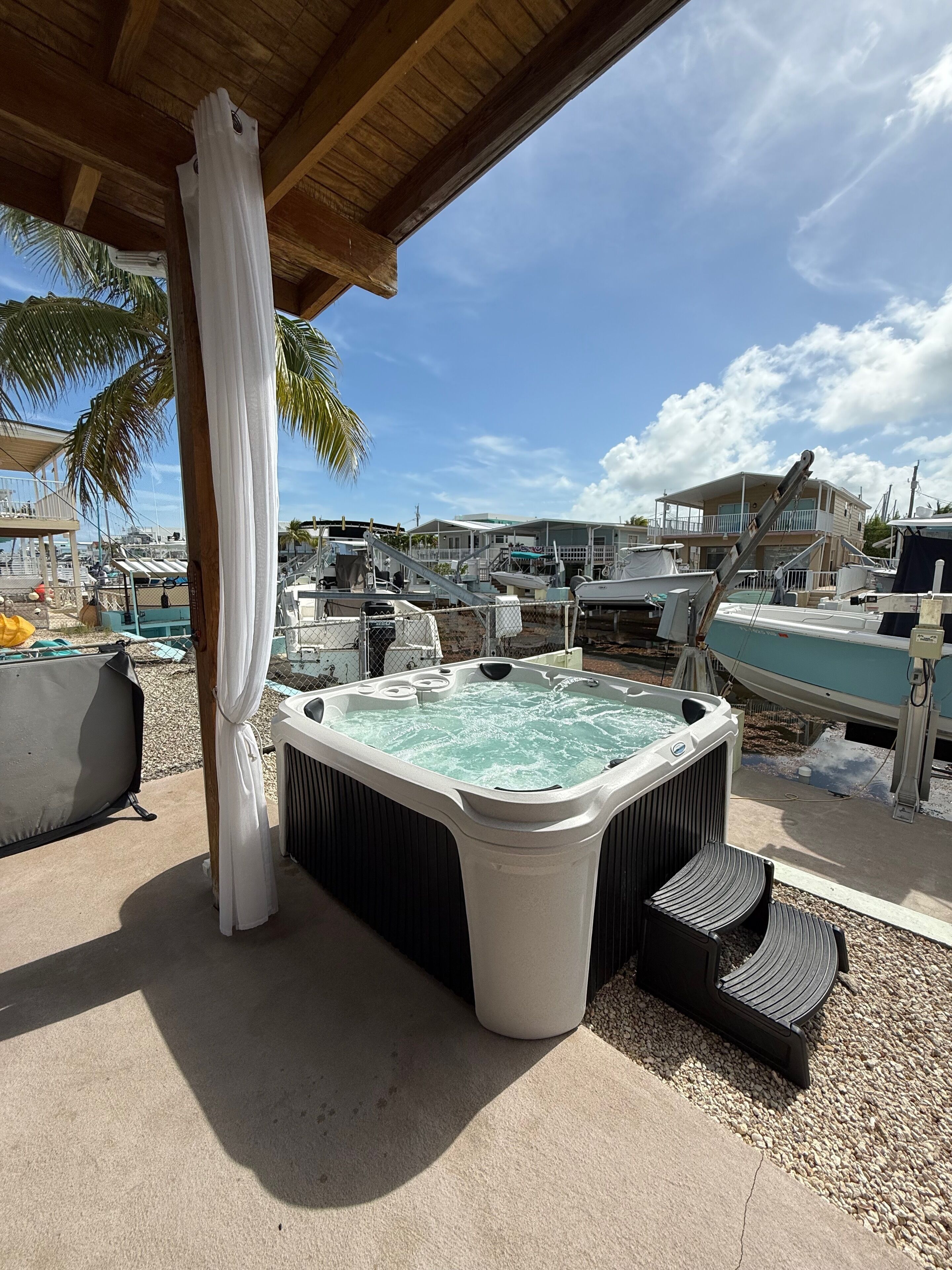 Un jacuzzi bajo una pérgola de madera con vista a barcos y un edificio al fondo.