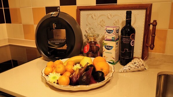 Uma bancada de cozinha com uma cafeteira, uma garrafa de vinho, uma caixa de chá Tetley e uma tigela de frutas.
